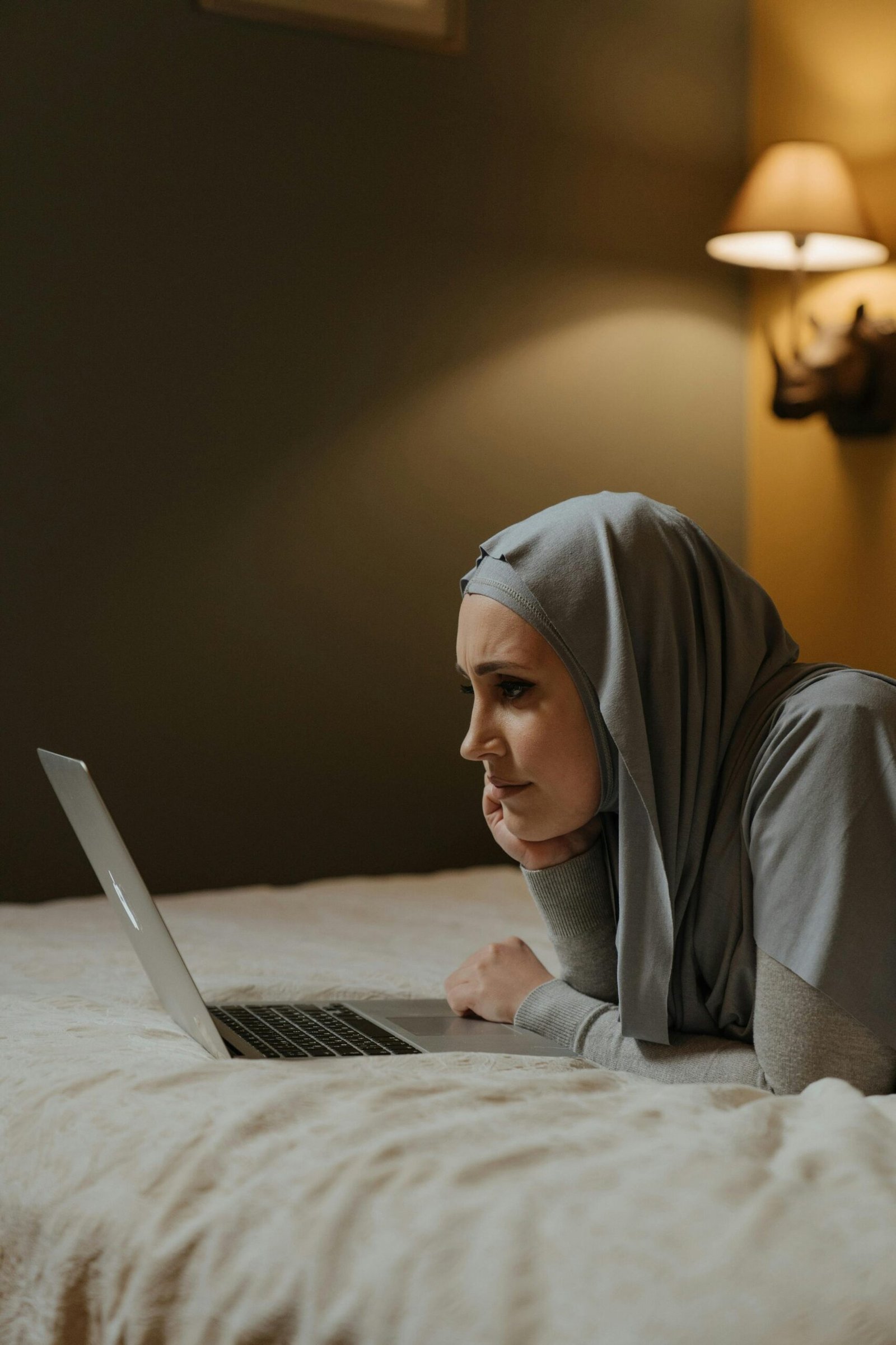 A Muslim woman in a hijab lies on a bed using a laptop, showcasing remote work in a cozy home setting.